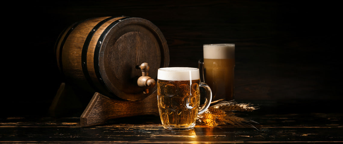 Rustikales Bierfass aus Holz mit Zapfhahn, daneben ein Glas frisch gezapftes, goldfarbenes Lagerbier mit Schaumkrone sowie ein Weizenbier im Hintergrund. Dekoriert mit Ähren, vor neutralem Hintergrund.
