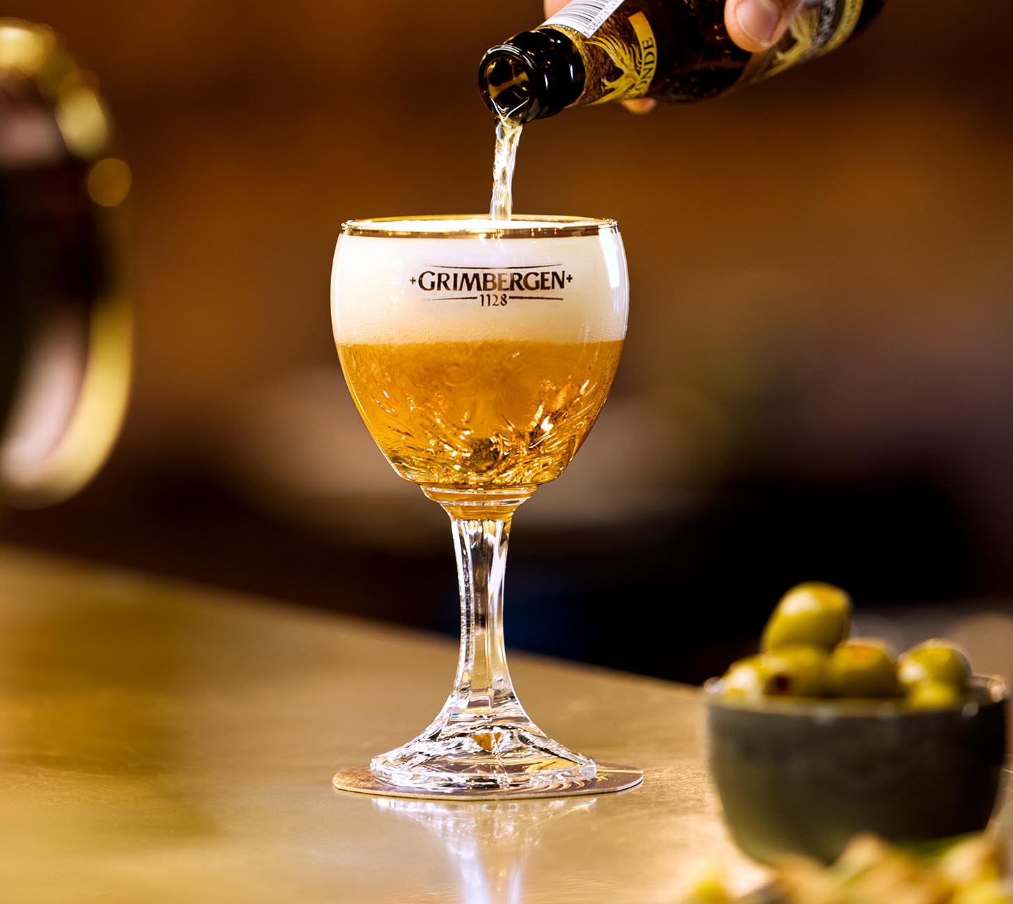 DE: Grimbergen Blonde Bier – goldgelbes Bier wird in ein Kelchglas mit Schaumkrone gegossen, warmes Stimmungsbild mit goldenem Licht.
FR: Bière Grimbergen Blonde – bière dorée versée dans un verre calice avec mousse, image d’ambiance chaleureuse à la lumière dorée.
IT: Birra Grimbergen Blonde – birra bionda dorata versata in un bicchiere a calice con schiuma, immagine d’atmosfera con luce dorata.