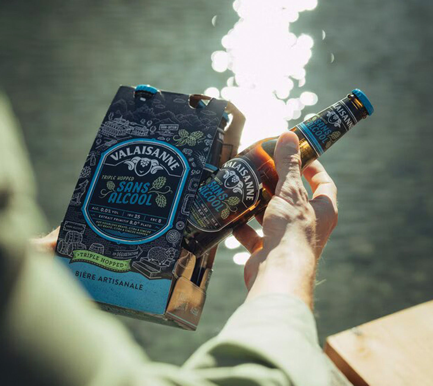 DE: Valaisanne Sans Alcool – Person hält Flasche und 4-Pack in natürlicher Outdoor-Szene am Wasser.
FR: Valaisanne Sans Alcool – une personne tient la bouteille et le pack dans une scène extérieure au bord de l’eau.
IT: Valaisanne Sans Alcool – persona con bottiglia e pack in una scena naturale vicino all’acqua.