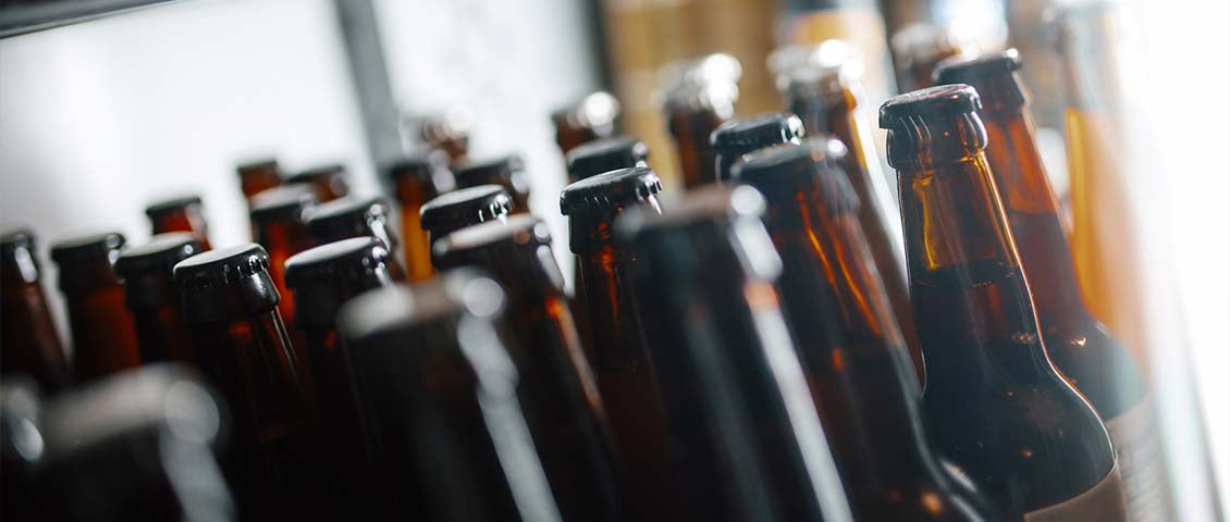 DE: Reihe dunkler Bierflaschen nebeneinander in Nahaufnahme FR: Rangée de bouteilles de bière sombres en gros plan côte à côte IT: Fila di bottiglie di birra scure in primo piano affiancate EN: Row of dark beer bottles side by side in close-up