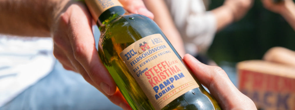 DE: Hand hält personalisierte Feldschlösschen Flasche mit Namen im Freien FR: Main tenant une bouteille Feldschlösschen personnalisée avec prénoms IT: Mano tiene bottiglia Feldschlösschen personalizzata con nomi all'aperto EN: Hand holding personalised Feldschlösschen bottle with names outdoors