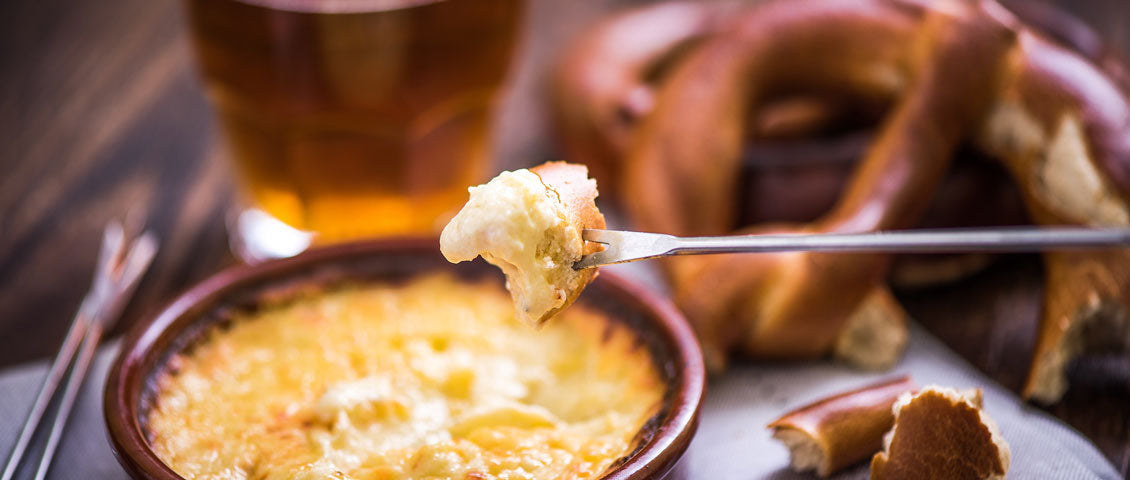 DE: Gabel taucht in cremiges Käsefondue in Keramiktopf mit Brezeln FR: Fourchette plongeant dans une fondue au fromage crémeuse avec bretzels IT: Forchetta immersa in fonduta di formaggio cremosa con bretzel EN: Fork dipping into creamy cheese fondue in ceramic pot with pretzels