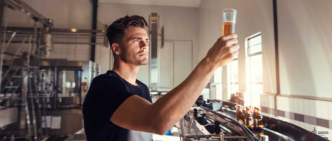 DE: Mann begutachtet Bierglas in moderner Brauerei vor Edelstahlkesseln FR: Homme examinant un verre de bière dans une brasserie moderne devant des cuves IT: Uomo esamina bicchiere di birra in birreria moderna davanti a serbatoi in acciaio EN: Man inspecting beer glass in modern brewery in front of stainless steel tanks