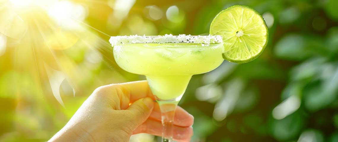 DE: Hand hält Margarita-Glas mit Salzrand und Limettenscheibe FR: Main tenant un verre de margarita avec bord salé et tranche de citron vert IT: Mano tiene bicchiere di margarita con bordo salato e fetta di lime EN: Hand holding margarita glass with salted rim and lime slice