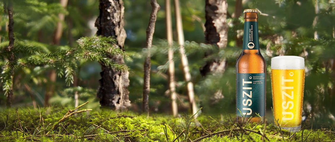 DE: USZIT Bier Glasflasche lehnt an Baumstamm im Wald FR: Bouteille en verre USZIT appuyée contre un tronc d'arbre en forêt IT: Bottiglia di vetro USZIT appoggiata a un tronco d'albero nel bosco EN: USZIT beer glass bottle leaning against tree trunk in forest