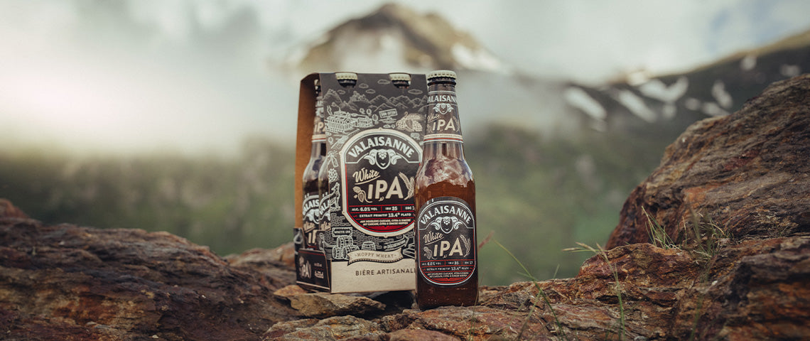 DE: Valaisanne White IPA Flaschen vor winterlichem Bergpanorama FR: Bouteilles Valaisanne White IPA devant panorama montagneux hivernal IT: Bottiglie Valaisanne White IPA davanti a panorama montano invernale EN: Valaisanne White IPA bottles in front of winter mountain panorama