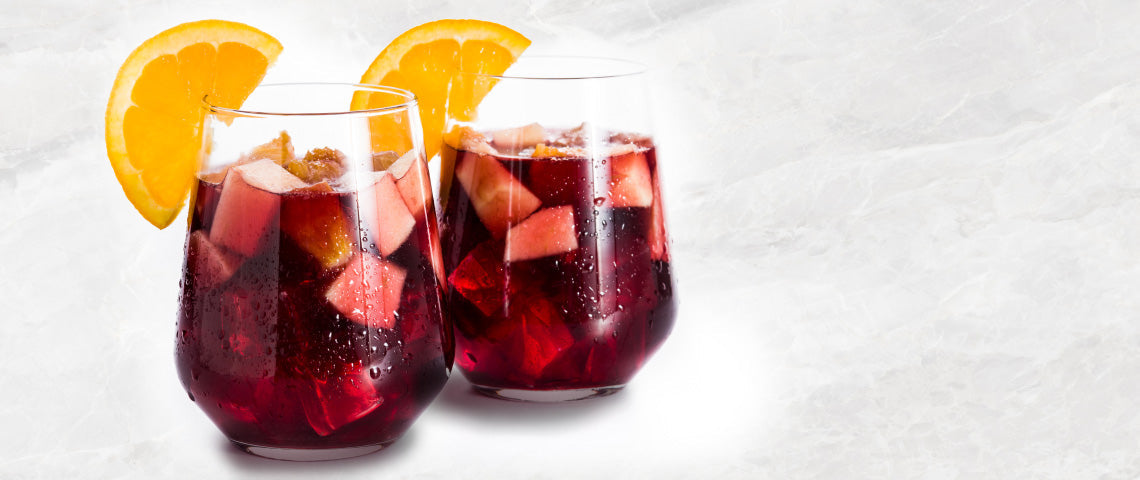 DE: Zwei Sangria-Gläser mit Orangenscheibe und Eiswürfeln auf hellem Hintergrund FR: Deux verres de sangria avec tranche d'orange et glaçons sur fond clair IT: Due bicchieri di sangria con fetta d'arancia e cubetti di ghiaccio su sfondo chiaro EN: Two sangria glasses with orange slice and ice cubes on light background