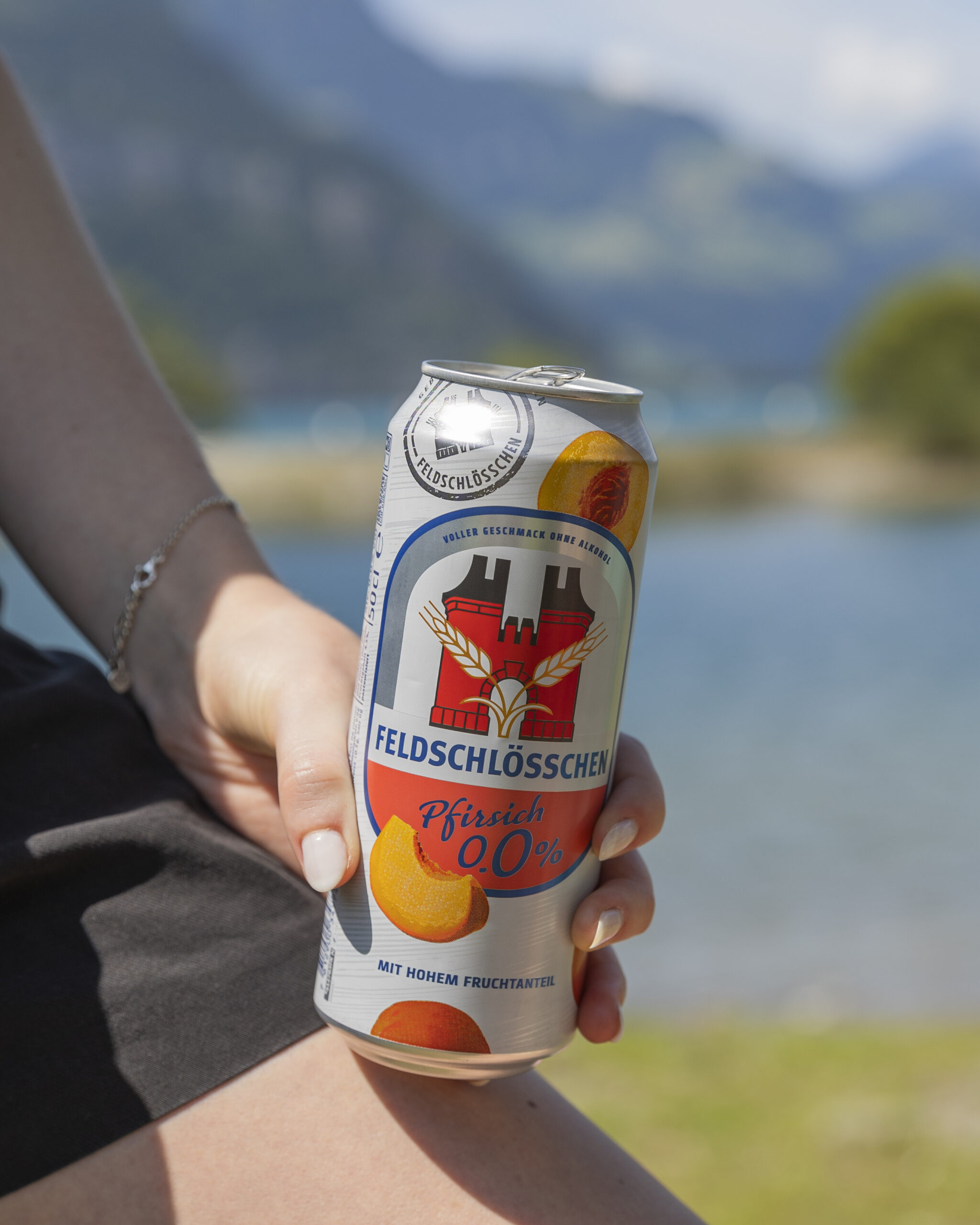 DE: Person hält Feldschlösschen Panaché 0.0% Pfirsich in der Hand, im Hintergrund ist ein See; FR: Une personne tient un Feldschlösschen Panaché 0.0% Pêche à la main, un lac en arrière-plan; IT: Una persona tiene in mano un Feldschlösschen Panaché 0.0% Pesca, sullo sfondo un lago; EN: A person holding a Feldschlösschen Panaché 0.0% Peach in hand, a lake in the background