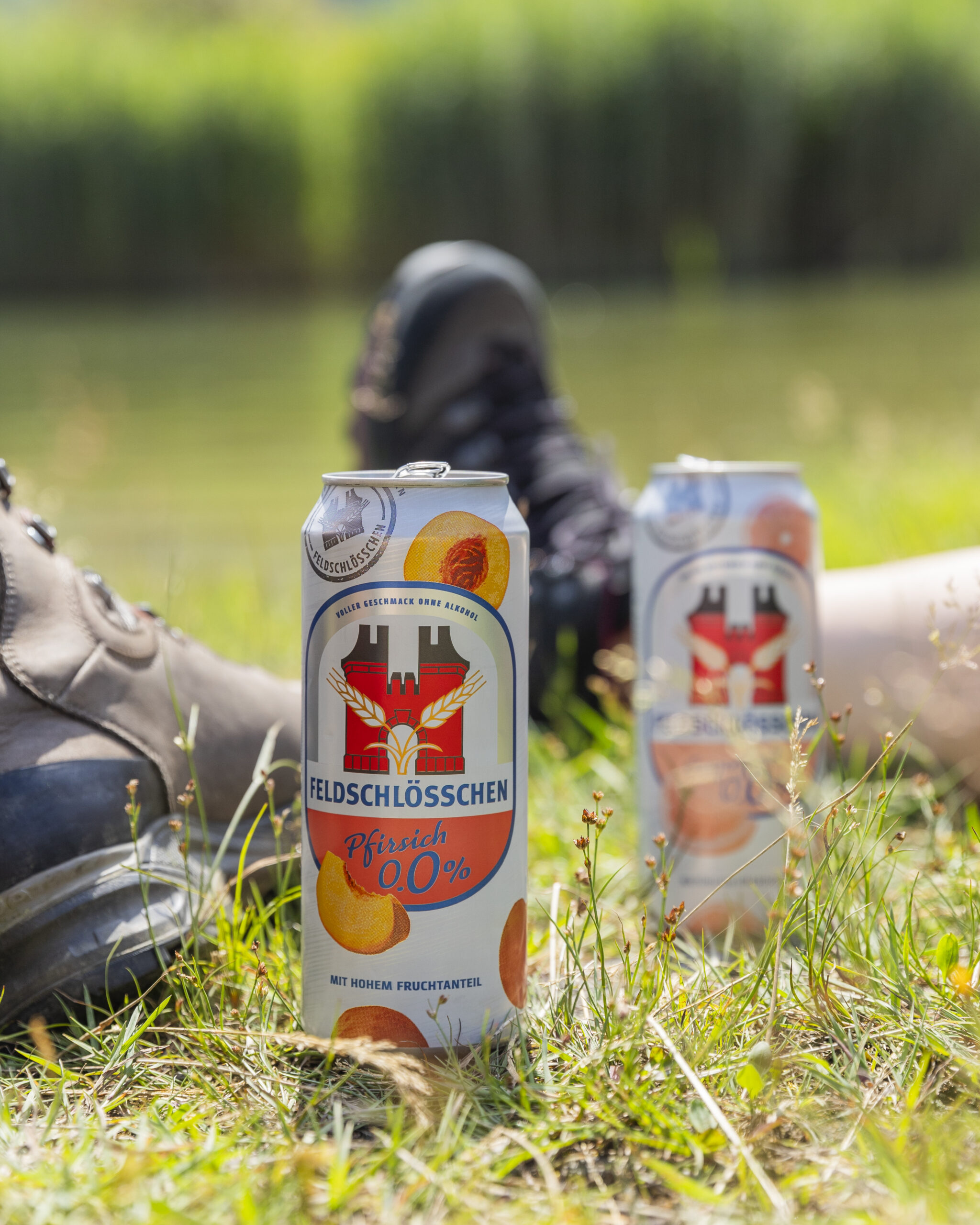 DE: Feldschlösschen Panaché 0.0% Pfirsich im Vordergrund, im Hintergrund Wanderschuhe und Feldschlösschen Panaché 0.0% Grapefruit;