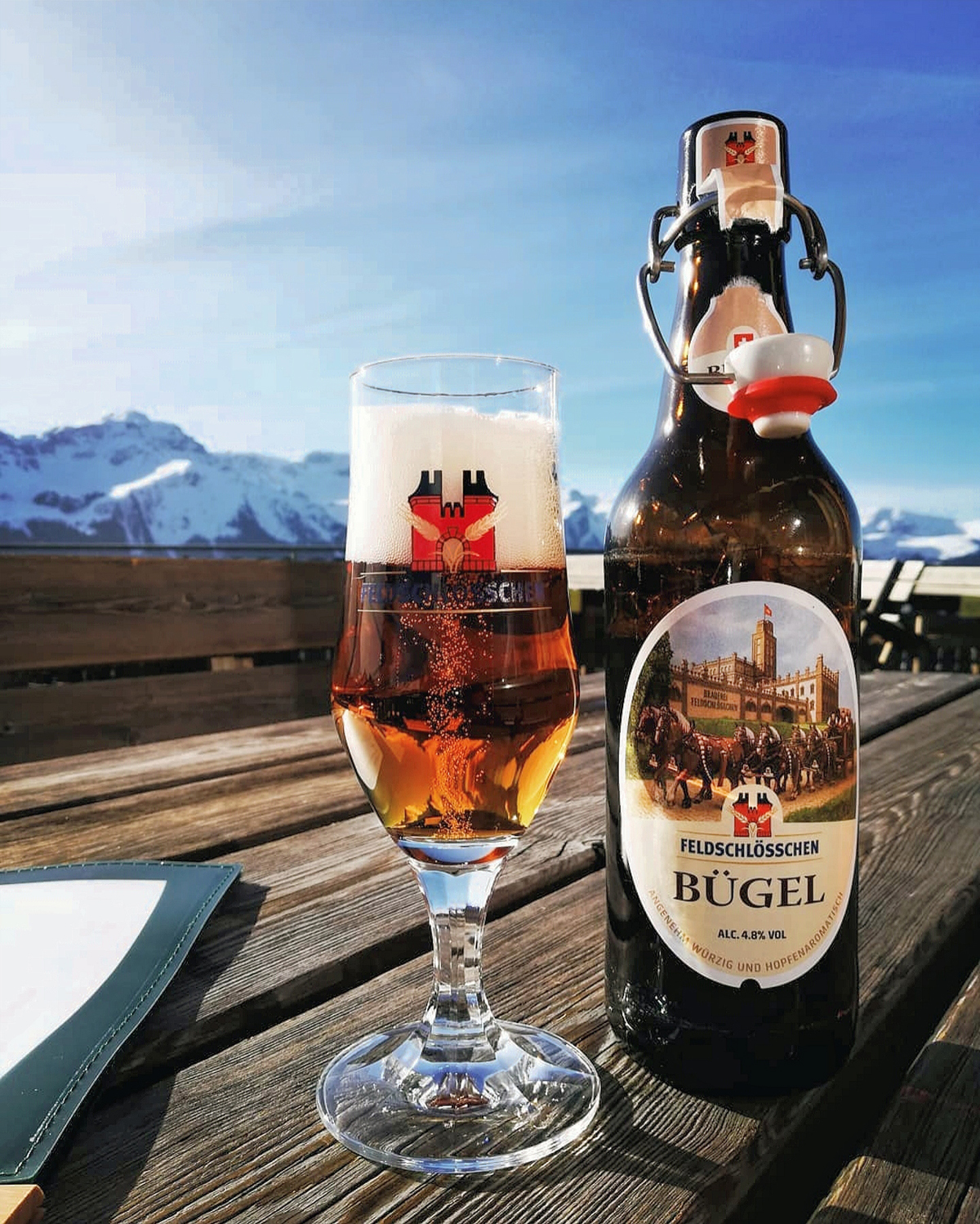 DE: Feldschlösschen Bügel Bier Flasche und eingeschenktes Bierglas mit schöner Bergaussicht; FR: Bouteille de bière Feldschlösschen à bouchon mécanique; verre de bière rempli avec une belle vue sur les montagnes; IT: Bottiglia di birra Feldschlösschen con tappo a leva; bicchiere di birra versata con una bella vista sulle montagne; EN: Feldschlösschen flip-top beer bottle; filled beer glass with a beautiful mountain view