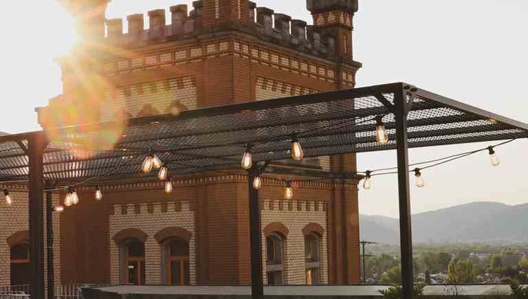 🇩🇪 Dachterrasse der Feldschlösschen Brauwelt Rheinfelden mit Lichterketten und Blick auf das Bierschloss bei Sonnenuntergang. 🇮🇹 Terrazza della Brauwelt Rheinfelden con luci decorative e vista sul castello della birra al tramonto. 🇫🇷 Terrasse de la Brauwelt Rheinfelden avec guirlandes lumineuses et vue sur le château de la bière au coucher du soleil. 🇬🇧 Rooftop terrace of the Feldschlösschen Brauwelt Rheinfelden with string lights and view of the beer castle at sunset.